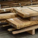 Thickness of kiln-dried unedged acacia boards stacked on a wooden pallet in a warehouse.