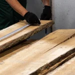 A worker wearing gloves measures the length of an unedged oak board, 52 mm, using a tape measure.