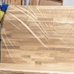 Worker holding a packaged oak furniture panel, showing the finger-joint pattern through the film.