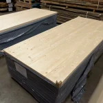Stack of solid-lameled pine furniture boards wrapped in protective film in a warehouse.
