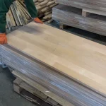 A warehouse worker in orange gloves holding a finger-jointed alder furniture board from a stack.